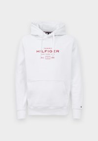 Hoodie blanc avec poche avant, capuche à cordon de serrage, et texte et logo rouges "Tommy Hilfiger New York City Est. 1985" sur la poitrine.