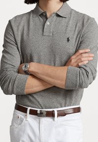 Graues Langarm-Polohemd mit klassischem Kragen, das ein kleines Logo auf der Brust trägt. Tragen mit weißen Hosen und einem braunen Gürtel.