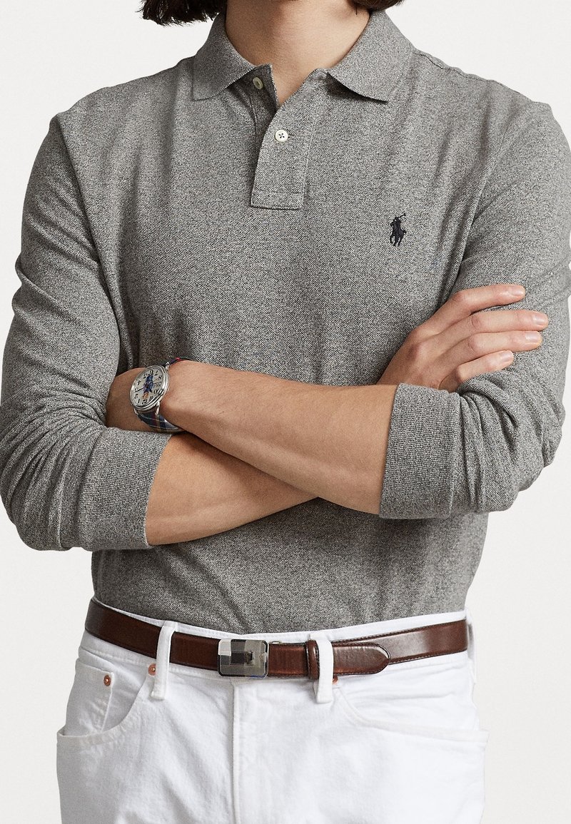 Graues Langarm-Polohemd mit klassischem Kragen, das ein kleines Logo auf der Brust trägt. Tragen mit weißen Hosen und einem braunen Gürtel.