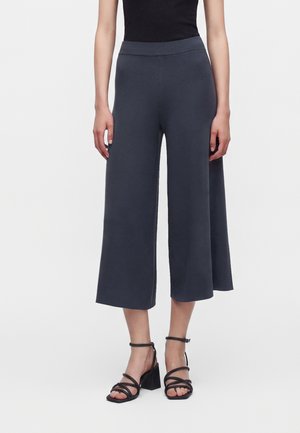 Woman wearing dark gray wide-leg cropped pants and black strappy high-heeled sandals standing against a plain background.