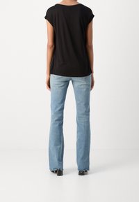 Kobieta stoi, ubrana w czarny krótkim rękawem top oraz jasnoniebieskie dżinsy bootcut z postrzępionymi brzegami i czarnych obcasach, odwrócona plecami.