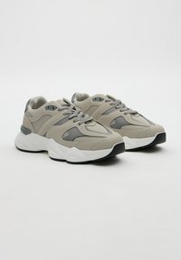 Un par de zapatillas deportivas beige y gris con suelas gruesas blancas y cordones, sobre un fondo blanco liso.