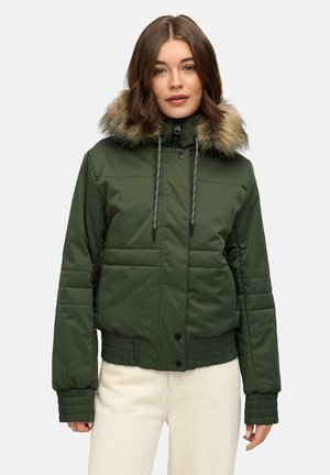 Vrouw in een groene gewatteerde jas met een met bont gevoerde capuchon, ritssluiting en drukknopen, geribbelde manchetten en crèmekleurige broek.