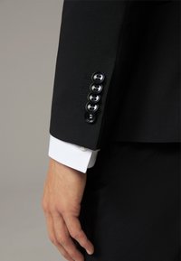 Schwarze maßgeschneiderte Anzugjacke mit fünf schwarzen Knöpfen und dezentem Stichdetail, kombiniert mit einem darunter sichtbaren weißen Hemdmanchet.