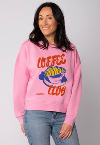 Roze katoenen sweatshirt met een grafisch ontwerp van een croissant in een kom, met de tekst "COFFEE CLUB" in vet oranje en blauw.
