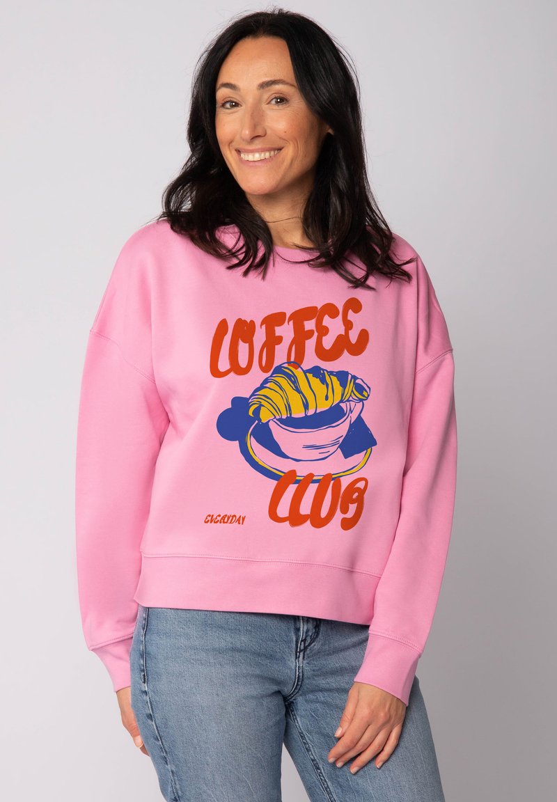 Roze katoenen sweatshirt met een grafisch ontwerp van een croissant in een kom, met de tekst "COFFEE CLUB" in vet oranje en blauw.