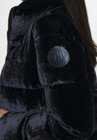 Giacca puffer di velluto blu navy con colletto alto, chiusura zip e una toppa rotonda con logo in rilievo sulla manica sinistra.