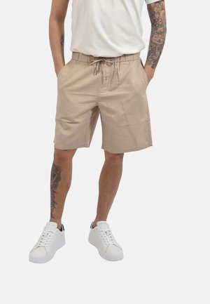 Uomo che indossa pantaloncini beige con coulisse, sneakers bianche e una maglietta bianca, con tatuaggi visibili su braccia e gambe.
