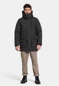 Giacca parka nera imbottita con cappuccio regolabile, due grandi tasche frontali e chiusura a zip. Il modello indossa pantaloni beige e stivali neri.