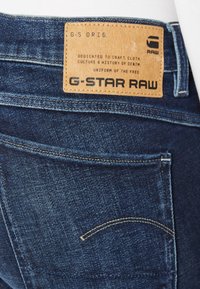 Mörkblå denimjeans med en texturerad yta, som har en beige läderetikett i bakre midjan med präglad logotyp.