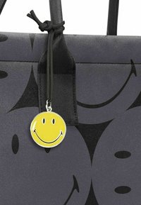Borsa tote in tessuto grigio con un motivo a facce sorridenti ripetuto. Presenta un'etichetta con un ciondolo a forma di faccina sorridente gialla attaccata da un cordino nero.