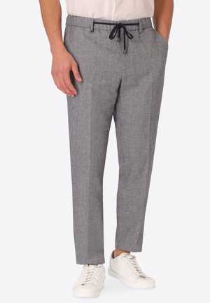 Uomo che indossa pantaloni grigi su misura con coulisse nera e sneaker bianche, in piedi con una mano in tasca e l'altra di fianco.