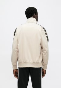 Bež sportska jakna s visokim ovratnikom, crnim bočnim prugama i rebrastim manšetama. Glatka tekstura, opušteni kroj i elastični struk.