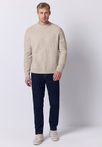 Beige gestrickter Pullover mit geripptem Rundhalsausschnitt und Saum, kombiniert mit dunkelblauen Jeans und hellen Sneakern; glatte Textur, lässiges Design.