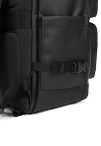 Mochila negra con superficie lisa, acentos de malla y correas ajustables. Cuenta con múltiples bolsillos y un cierre de hebilla resistente.