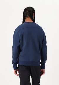 Laivansinis nešiojamas megztinis, pagamintas iš minkštos medžiagos, su apvalia kaklo linija, nukritusiomis pečių siūlėmis ir guminiu praktišku užbaigimu. Lygus tekstūra su minimaliais detalėmis.