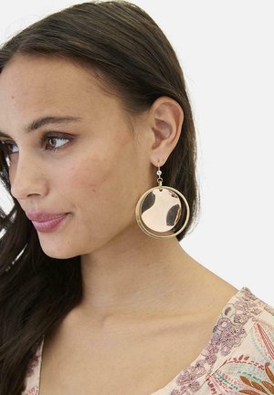 Joven mujer con cabello largo y oscuro lleva grandes pendientes redondos dorados de doble disco y una blusa con estampado, mirando ligeramente hacia la izquierda.