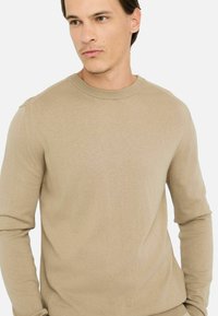 Beige kötött pulóver, kerek nyakkivágással, hosszú ujjakkal és sima textúrával, amely a testhez simuló megjelenésre lett tervezve.