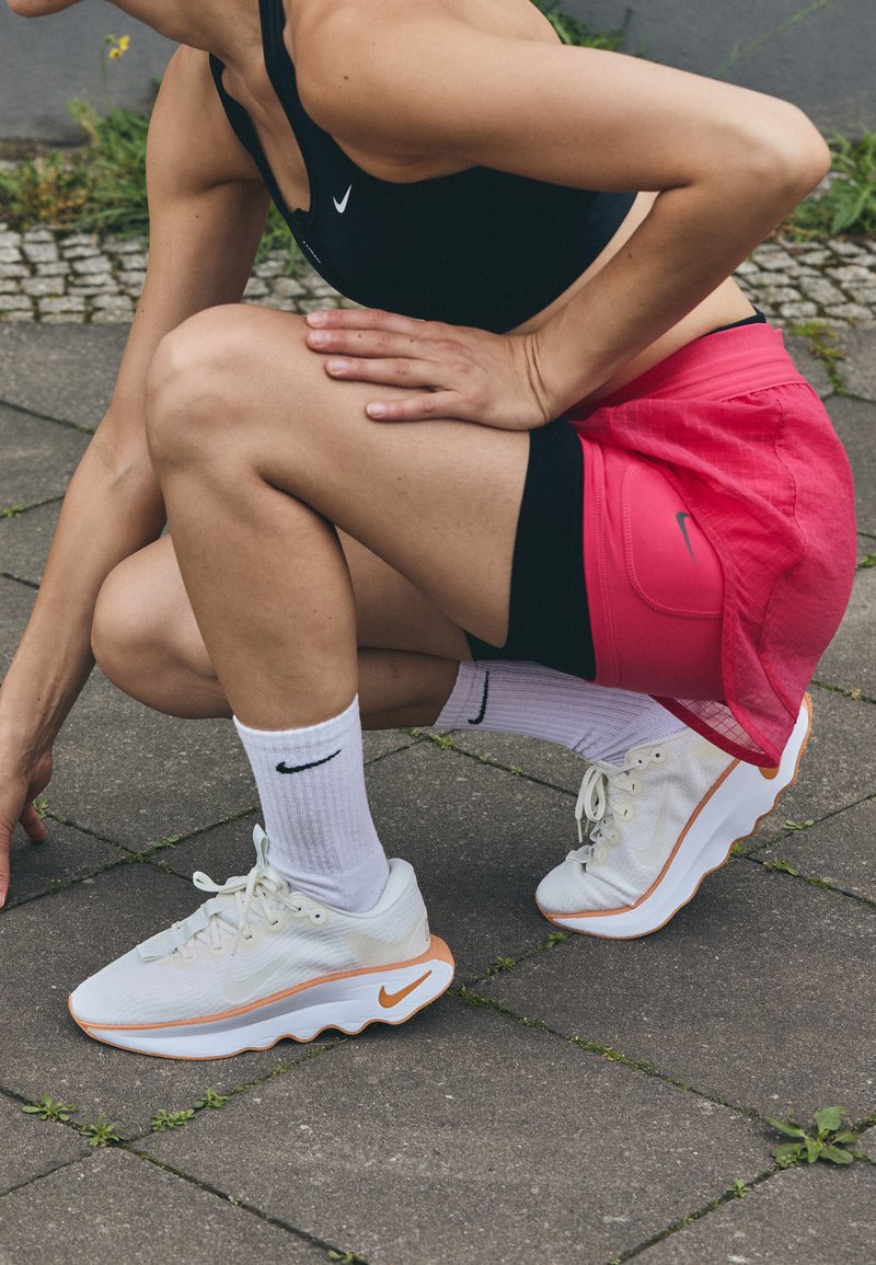 Baskets de sport Nike blanches avec des accents orange, accompagnées de chaussettes blanches, de shorts noirs et d'une jupe rose, sur un fond de pavé.