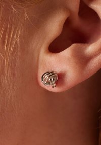 Boucle d'oreille en métal en forme de nœud, présentant des boucles entrelacées et une finition polie, portée sur un lobe, avec une peau claire en arrière-plan.