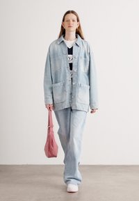 Veste oversize en denim bleu clair avec poches, associée à un jean assorti et une chemise graphique noir et blanc. Tient un sac à épaule rose.