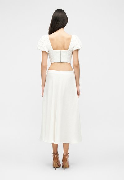 Femme aux longs cheveux foncés portant un crop top blanc à manches bouffantes avec fermeture éclair dans le dos et une jupe midi blanche fluide, debout de dos.