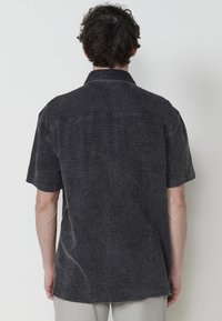 Camisa de manga corta en tejido negro texturizado con un patrón sutil, cuenta con un collar y un dobladillo recto. La vista trasera muestra un detalle minimalista.