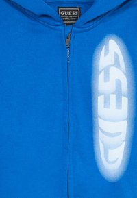 Hoodie azul com fecho éclair feito de tecido macio, apresentando um proeminente logotipo branco "GUESS" com um efeito de gradiente no lado direito.