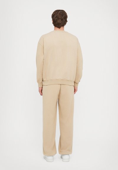Beige sweatshirt og matchende løstsittende bukser, blødt stof, ribbede manchetter og kant, minimalistisk design, ensfarvet bagside, hvide fodtøj.