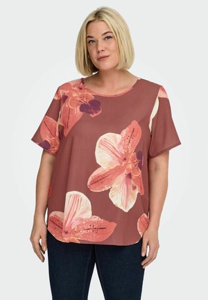 Kvinde med blond bob-paryk iført en løs, kortærmet top med store pink og ferskenfarvede blomsterprint samt mørke bukser.