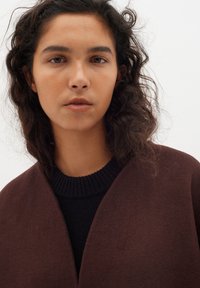 Rudas vilnos mišinio viršutinė apranga su giliu V formos iškirpimu, uždėta ant tekstūrinio tamsaus mezginio megztinio. Natūralūs garbanoti plaukai rėmina veidą.