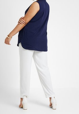 Person wearing sleeveless navy blue blouse, white pants, and white high-heeled shoes, standing with arms crossed and back turned.