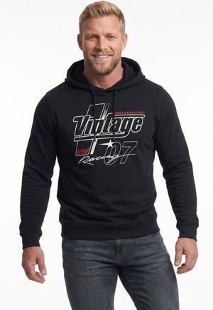Uśmiechnięty mężczyzna z krótkimi blond włosami, ubrany w czarną bluzę z kapturem z napisem "Vintage 07 Racing" oraz niebieskie dżinsy, stojący na białym tle.