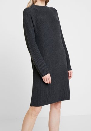 Femme portant une robe en maille côtelée gris foncé à manches longues, debout devant un fond clair uni.