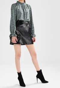 Blouse verte à pois noirs avec un col montant, associée à une jupe en fausse cuir noir brillante. Des bottines noires complètent la tenue.
