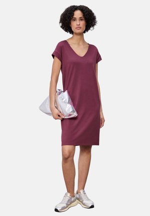 Frau mit kurzen lockigen Haaren, die ein knieumspielendes bordeauxrotes Kleid, silberne Turnschuhe trägt und eine metallisch silberne Clutch in der Hand hält.