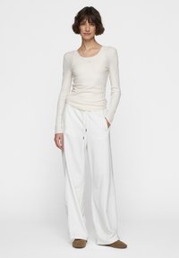 Pantaloni bianchi a gamba larga con vita regolabile abbinati a un top fitted a maniche lunghe in caldo beige a costine. La modella è in piedi su uno sfondo neutro.