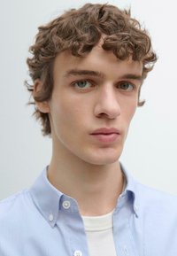 Jonge man met krullend bruin haar en groene ogen, die een lichtblauw gestreept overhemd draagt over een wit T-shirt, met een neutrale uitdrukking.