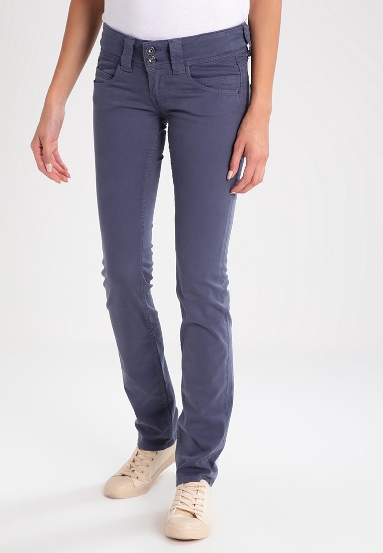 Femme portant un pantalon slim bleu foncé et des baskets crème, debout sur un fond blanc uni avec les mains détendues le long du corps.