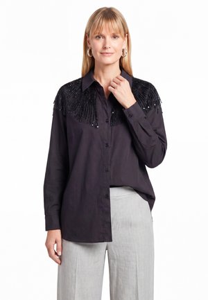 Blonde vrouw in zwart, met knopen gesloten overhemd met kralenfranje op de schouders en lichtgrijze broek, staand tegen een witte achtergrond.