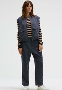 Ärmellose dunkelblaue Weste mit zwei Fronttaschen, kombiniert mit einem gestreiften Langarmshirt und locker sitzenden dunklen Hosen, weiße Sneakers.