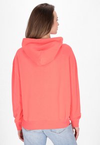 Korálová mikina s klokaní kapsou, oversize střih a vroubkované manžety. Hladký materiál, vybavená kapucí se stahovací šňůrkou. Stylizováno s světle modrými džíny.