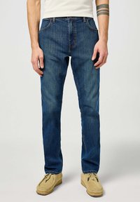 Jeans de denim azul con corte recto, cinco bolsillos y costuras naranjas en contraste. Llevados con zapatos de estilo mocasín beige.