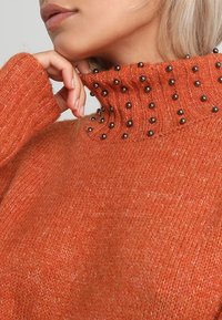 Maglione arancione lavorato a maglia con un alto colletto a costine e dettagli di perline nere. La texture sembra morbida e leggermente pelosa.