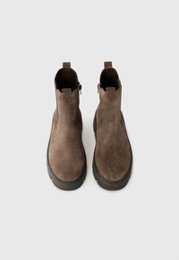 Bottines en daim marron avec une texture lisse, bout rond, semelle en caoutchouc noir épaisse et fermetures éclair sur les côtés pour un enfilage facile.