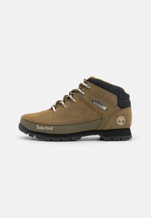 Botte de randonnée en cuir marron avec col rembourré noir, œillets métalliques, lacets à motifs, semelle robuste et logos Timberland sur le côté et le talon.