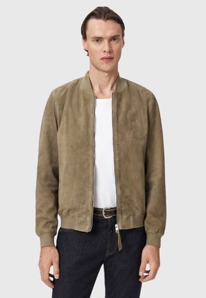 Ein Mann steht, trägt eine hellbraune Wildlederjacke über einem weißen Hemd und einer dunklen Jeans, und blickt nach vorne vor einem schlichten, hellgrauen Hintergrund.