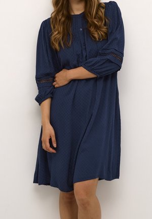 Donna che indossa un vestito blu navy al ginocchio con maniche a tre quarti e dettaglio in pizzo, in piedi davanti a uno sfondo chiaro e uniforme.