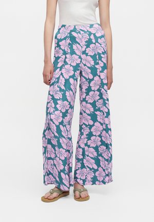 Personne portant un pantalon large couleur sarcelle avec un grand imprimé floral rose et des sandales beiges, debout devant un fond blanc uni.
