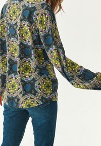 Camicia a maniche lunghe con un vivace motivo floreale astratto nei colori verde, blu e rosa, realizzata in tessuto leggero, abbinata a jeans blu.